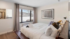 A hotel room with a large bed, a bedside table with a lamp, a framed artwork on the wall, and a window with curtains. - Spacious Condo with Lake and Mountain View (Mont-Tremblant)
