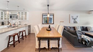 A modern kitchen with white cabinets, a wooden dining table, and bar stools. There is a gray sofa and a coffee table in the living area. - Spacious Condo with Lake and Mountain View (Mont-Tremblant)