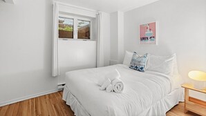 A bedroom with a bed, a window, a wall-mounted picture, and a bedside table with a lamp.