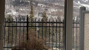A view from a window with a metal railing, overlooking a snowy landscape with mountains and a town below.