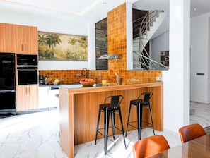 A modern kitchen with wooden cabinets, a central island with stools, a stone backsplash, and a staircase. - Rooftop Sea View Villa Near Beach (Essaouira)
