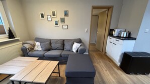 Ein Wohnzimmer mit einer Couch, einem Holztisch und einer Küchenzeile mit Schränken und Geräten.