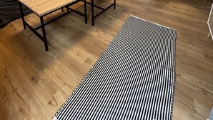 Ein modernes Wohnzimmer mit einer Couch, einem Holztisch und einem Streifen Teppich. Im Hintergrund ist eine Küche mit Essbereich zu sehen.