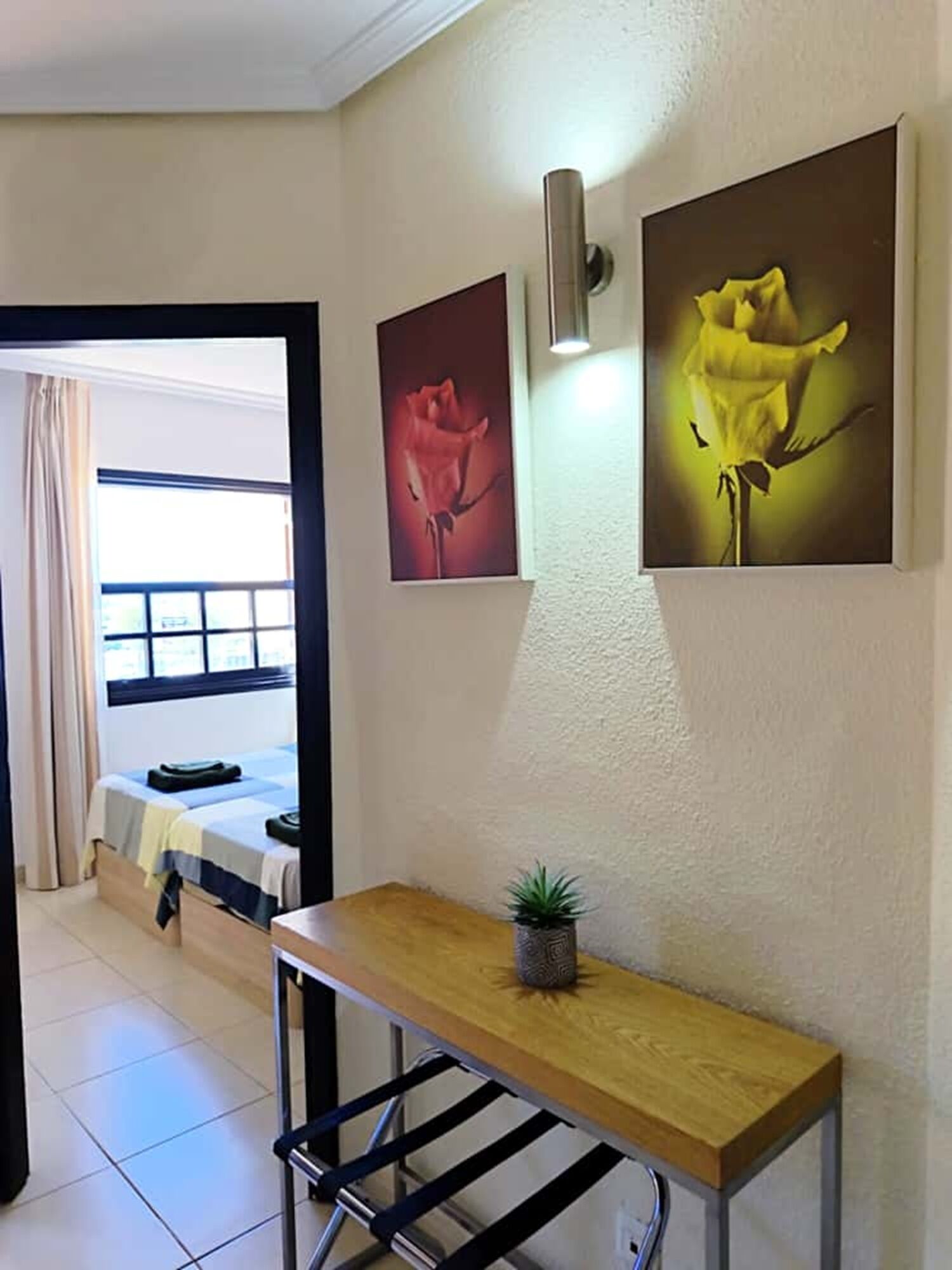 Habitación de hotel con cama, una mesa de madera con planta en maceta y dos cuadros en la pared.