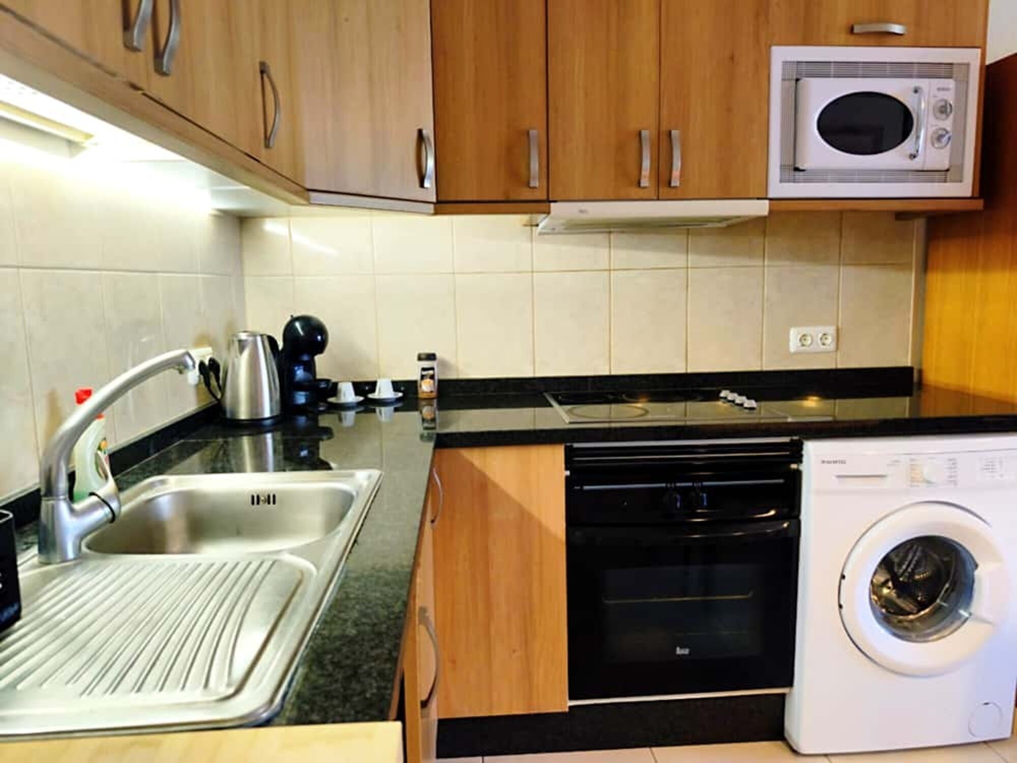 Una cocina compacta con armarios de madera, microondas integrado, lavavajillas y lavadora.