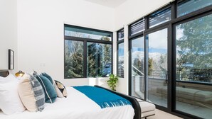 A modern bedroom with a large bed, multiple pillows, a bedside lamp, and a view of a snowy landscape through large windows. - 5 Min to Bald Mountain | Sawtooth Views (Ketchum)