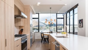 A modern kitchen with wooden cabinets, a large island with a sink, and a view of mountains through large windows. - 5 Min to Bald Mountain | Sawtooth Views (Ketchum)