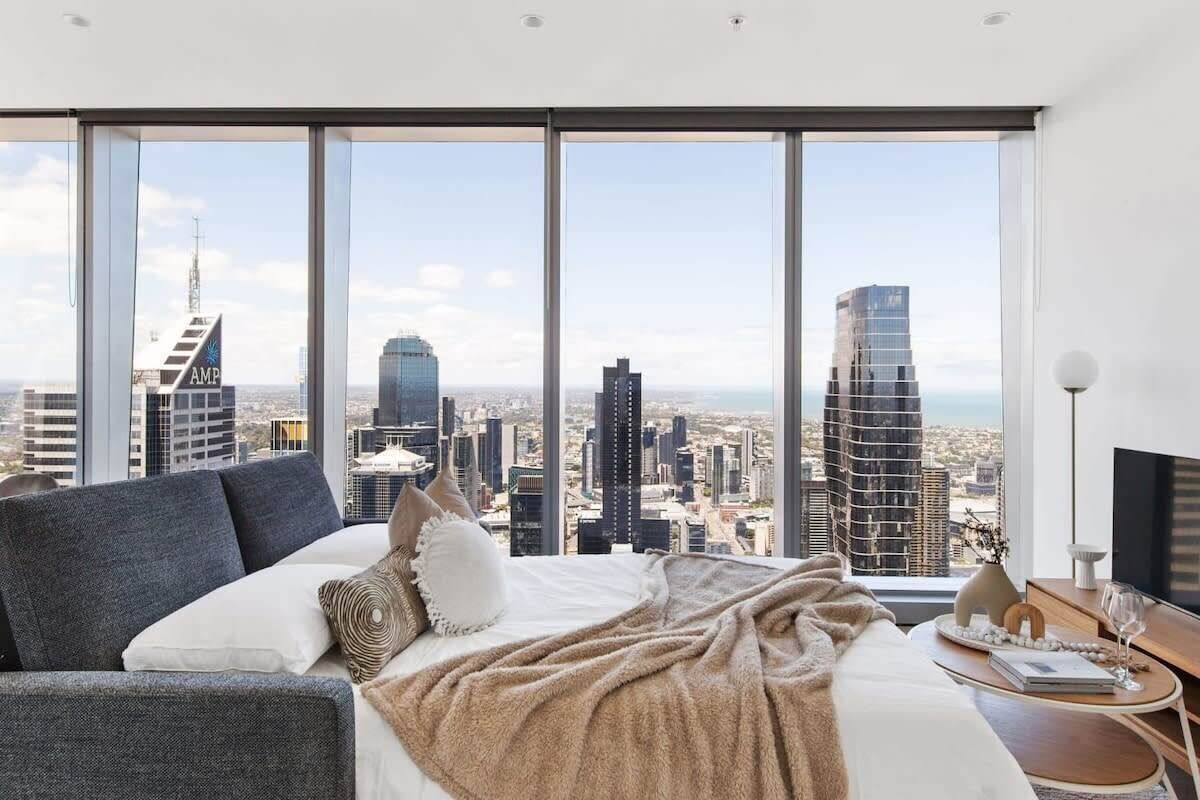 A modern hotel room with a large bed, a sofa, and a city view through floor-to-ceiling windows.