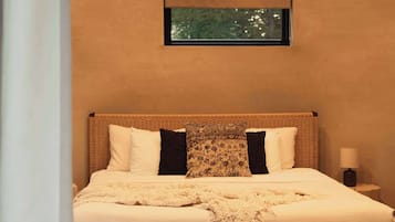 Een slaapkamer met een groot bed, twee nachtkastjes, een geweven bankje en een ingelijst schilderij aan de muur.