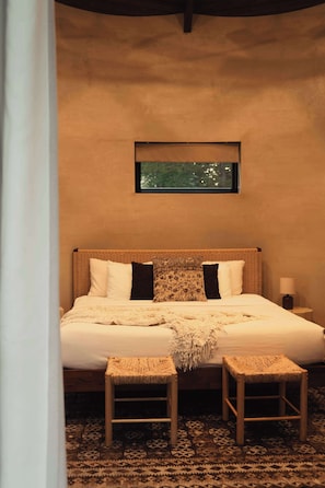 Un dormitorio con una cama grande, dos mesitas de noche, un banco tejido y un cuadro enmarcado en la pared.
