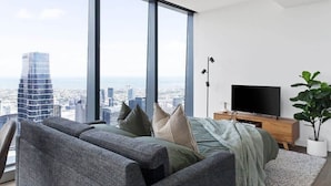 A modern living room with a large window, a sofa, a TV, and a plant. - A 2BR Apt with Amazing City Views, FREE Parking (Melbourne)
