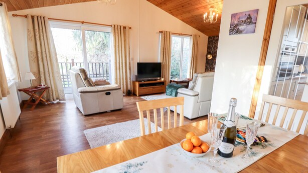 A spacious living area with a wooden floor, a dining table with a placemat, a sofa, a TV, and a window with curtains. - Kantankye Lodge (Skegness)