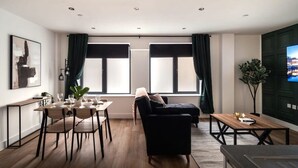 Ein modernes Wohnzimmer mit Sofa, Esstisch und Stühlen. Große Fenster mit Vorhängen, eine Pflanze und ein Flachbildfernseher auf einem Standfuß.