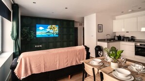 Eine moderne Küche mit weißen Schränken, eine Essecke mit Tisch für zwei Personen und ein Fernseher an einer grünen Wandpaneele.