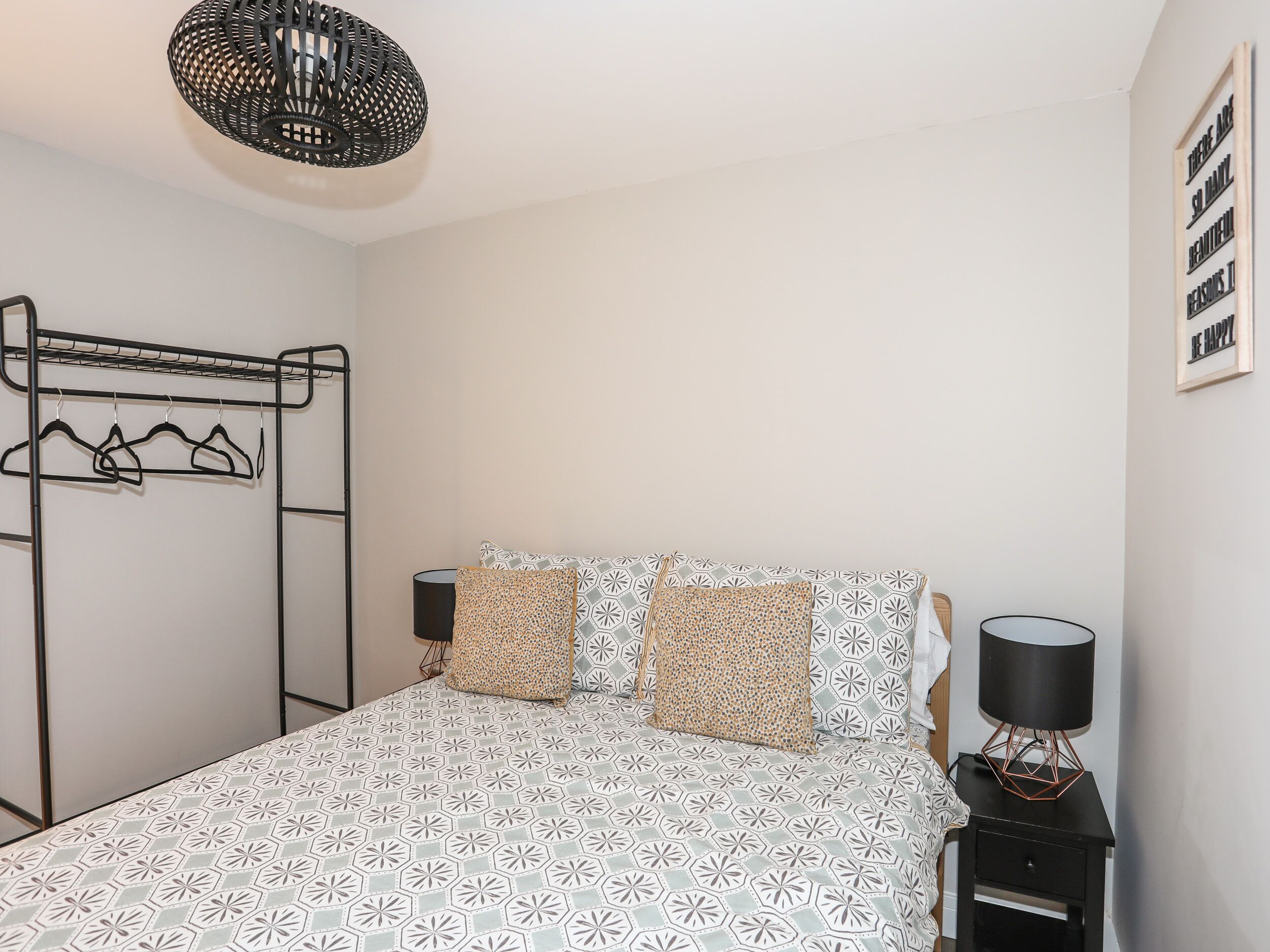 A bedroom with a patterned bedspread, a black metal wardrobe, a bedside table with a lamp, and a wall-mounted light fixture.