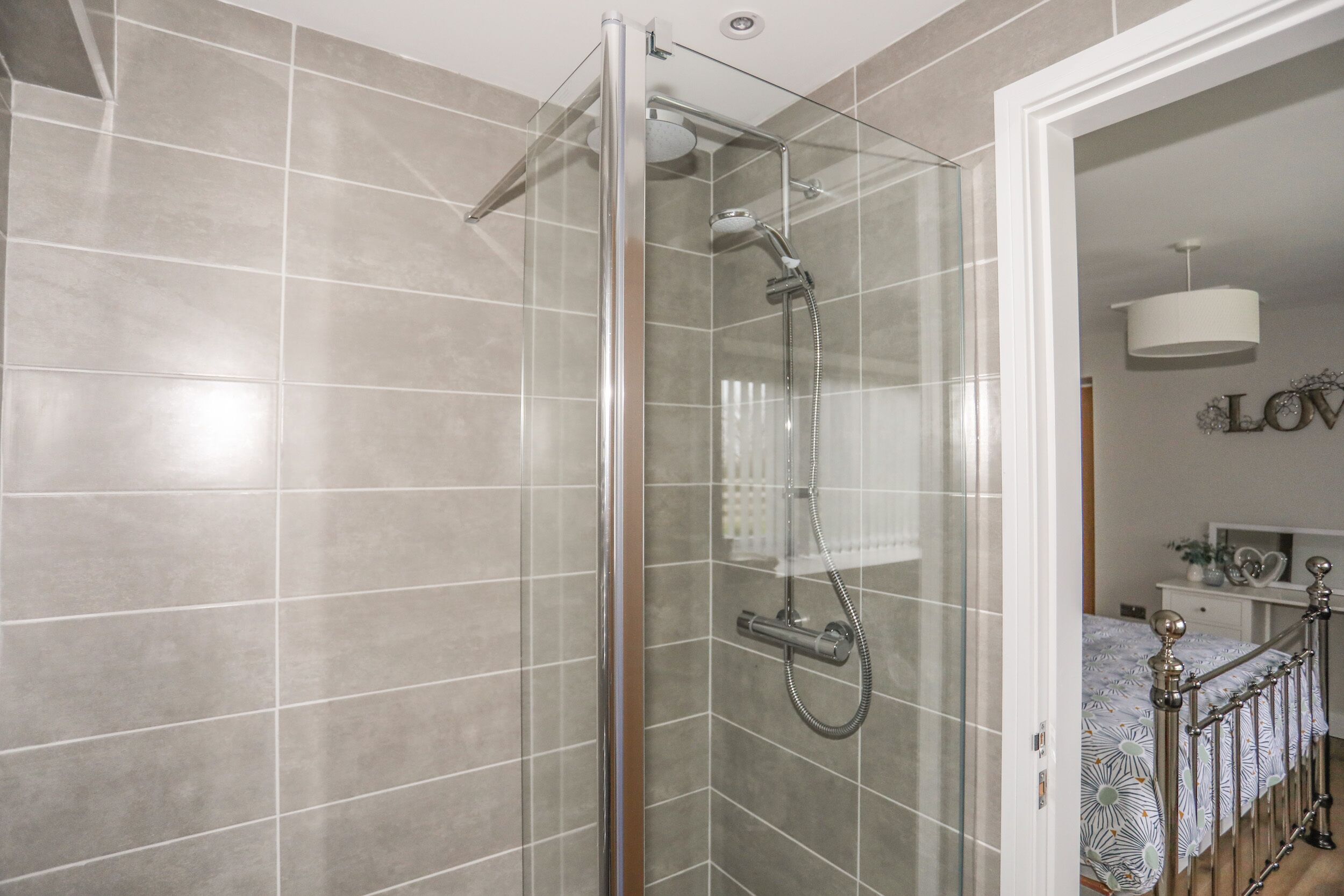 A modern bathroom with a glass-enclosed shower, tiled walls, and a bed with a patterned bedspread in the adjacent room.