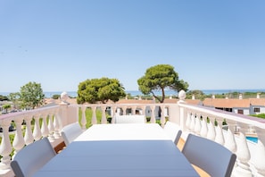 A terrace with a table and chairs, overlooking a coastal area with trees and buildings.
