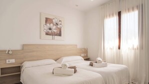 Una habitación de hotel con dos camas, un cabecero de madera, un cuadro de flores y una ventana con cortinas ligeras.