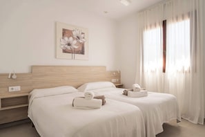 A hotel room with two beds, a wooden headboard, a painting of flowers, and a window with sheer curtains.