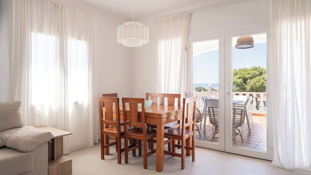 Un comedor con mesa y sillas de madera, un sofá y vistas a un patio exterior a través de un ventanal.