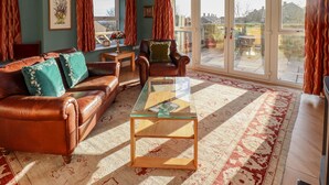 Ein Wohnzimmer mit einer Couch, Sesseln, einem Glastisch und Blick ins Freie durch große Glasüren.