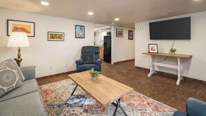 Un salon avec un canapé, une table basse, un fauteuil et un téléviseur. Les murs sont ornés de tableaux encadrés et un tapis recouvre le sol.