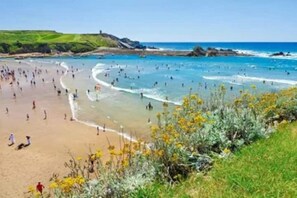 A beach with people swimming and walking along the shore, surrounded by greenery and a clear sky.