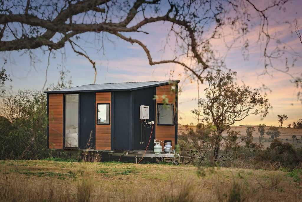 Une cabane moderne, de plain-pied, avec une façade sombre et des éléments décoratifs en bois, située dans un paysage naturel composé d’arbres et de vastes champs.
