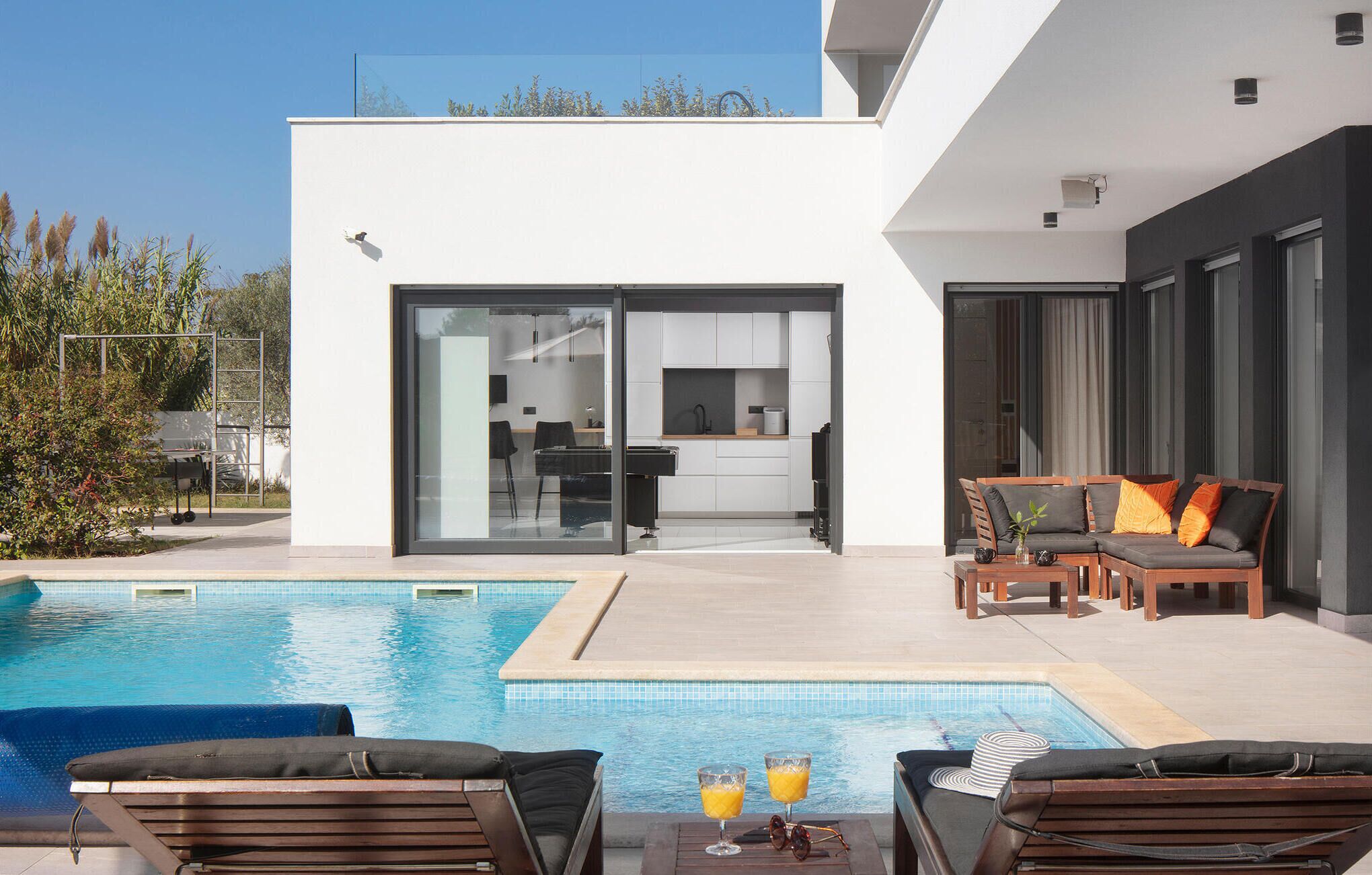 A modern outdoor pool area with lounge chairs and a dining table, a kitchen with sliding doors, and a view of the surrounding landscape.