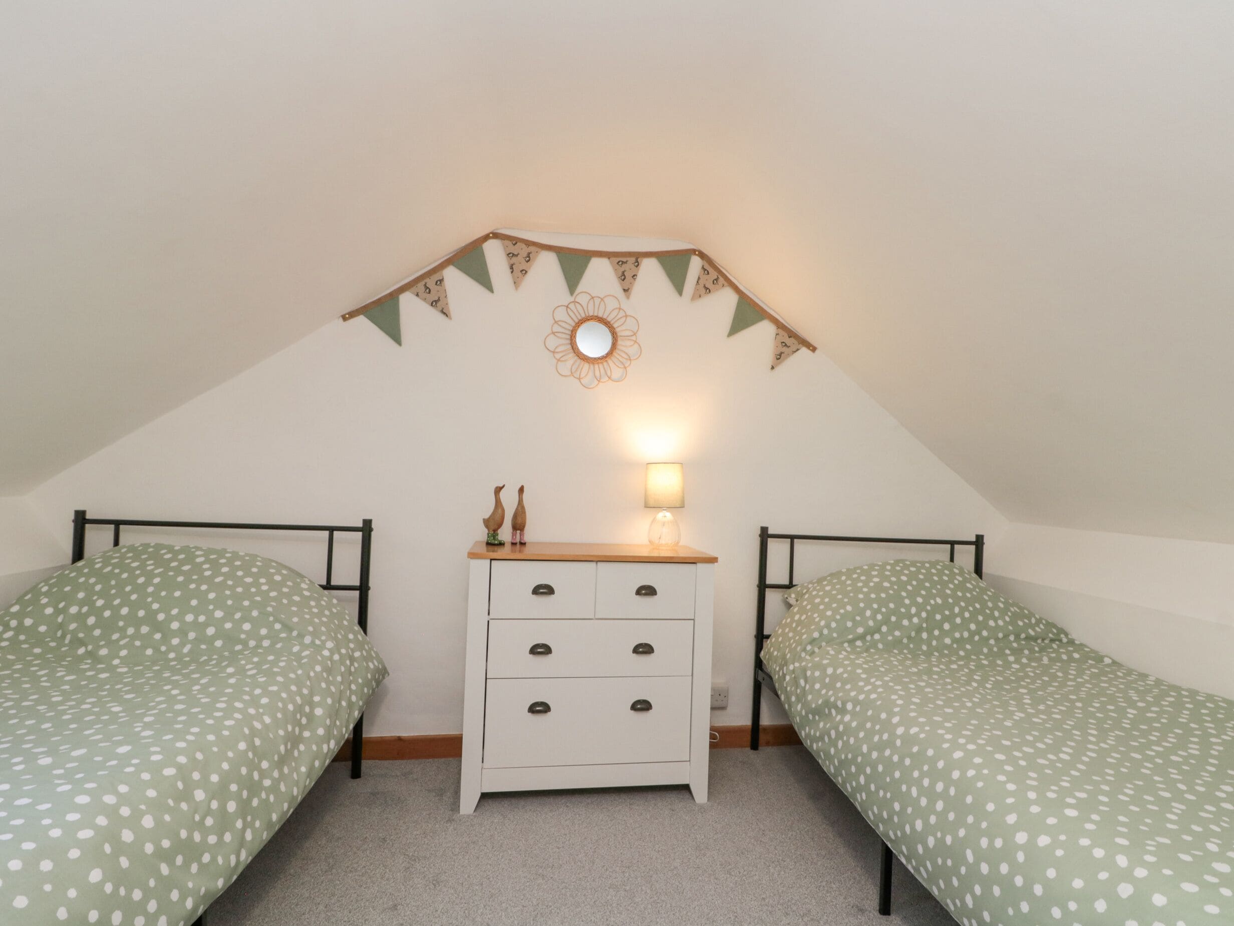 Una habitación pequeña y acogedora con dos camas individuales, un aparador blanco con cajones, un espejo y una lámpara.