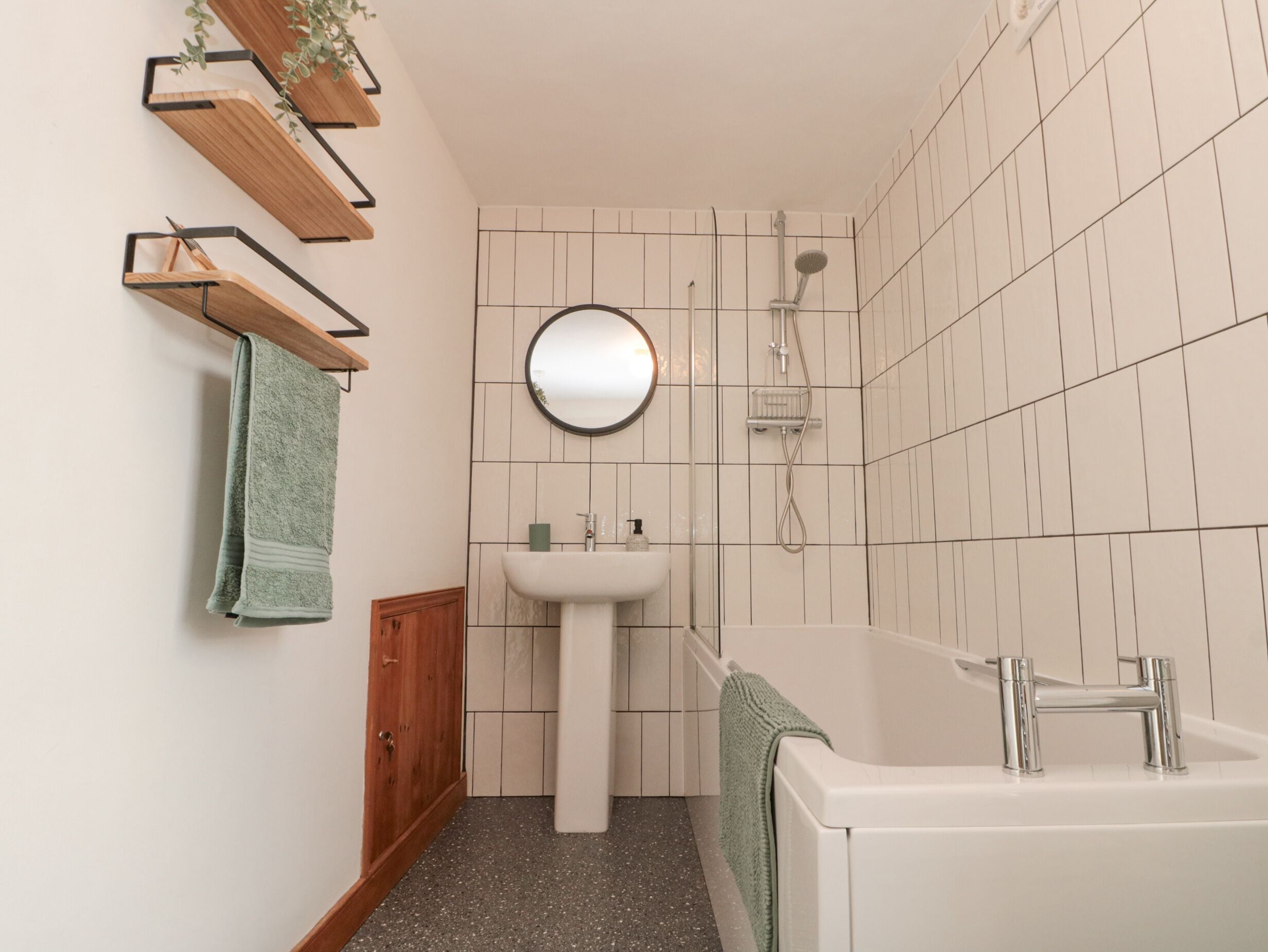 A bathroom with a white sink, a round mirror, a shower, and a bathtub. There are wooden shelves above the sink and a green towel hanging on the wall.