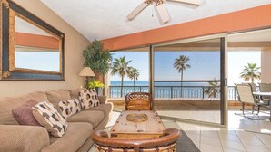 A living room with a sofa, a wooden coffee table, and a ceiling fan. There is a large window with a view of palm trees and the ocean.
