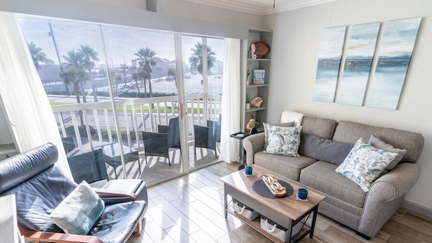 A living room with a grey sofa, a coffee table, and a balcony with a view of palm trees and a road. - Paradise Found! Stop Looking! (Galveston)