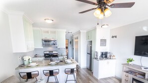 A modern kitchen with white cabinets, a central island with bar stools, a ceiling fan, and a flat-screen TV. - Paradise Found! Stop Looking! (Galveston)