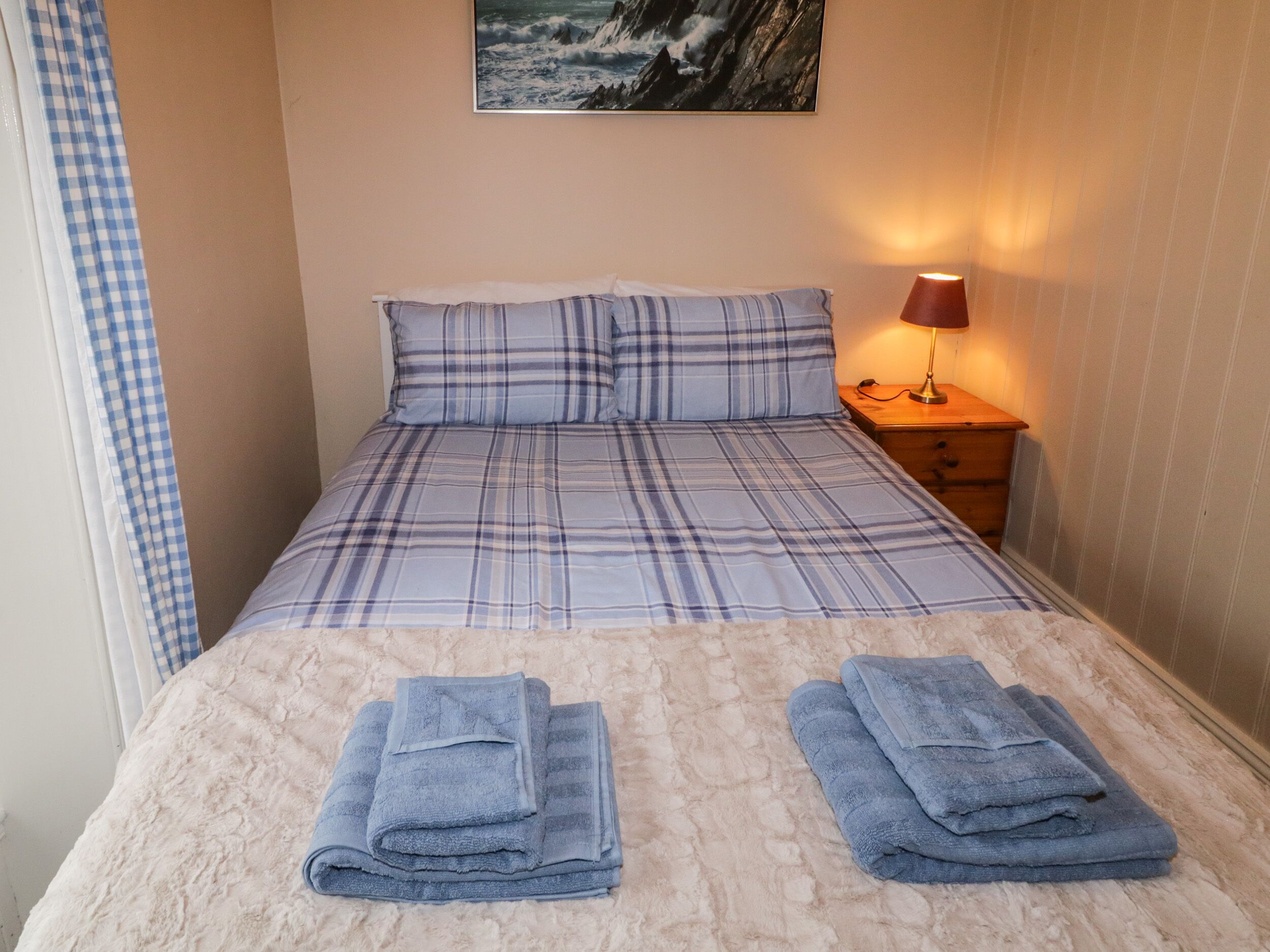 A neatly made bed with a plaid bedspread and pillows, a bedside lamp, and two folded towels on the floor.