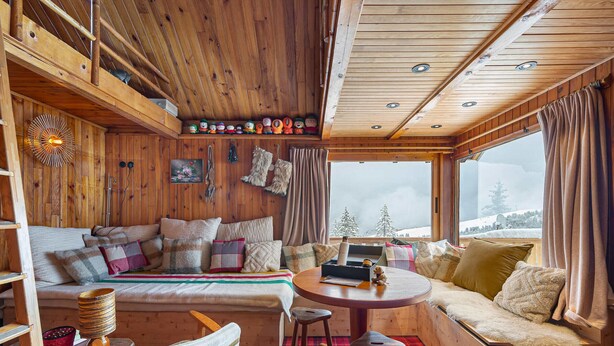 A cozy cabin interior with a wooden ceiling, a sofa with multiple cushions, a small table with a laptop, and a window with a snowy view.