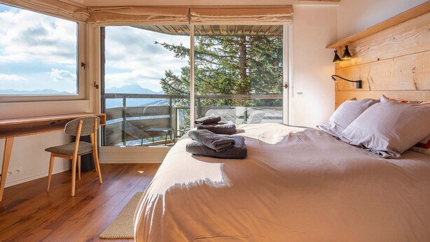 Ein Schlafzimmer mit Holzboden, einem Bett mit Kissen, einem Sessel und Blick auf Bäume durch eine Glastür.
