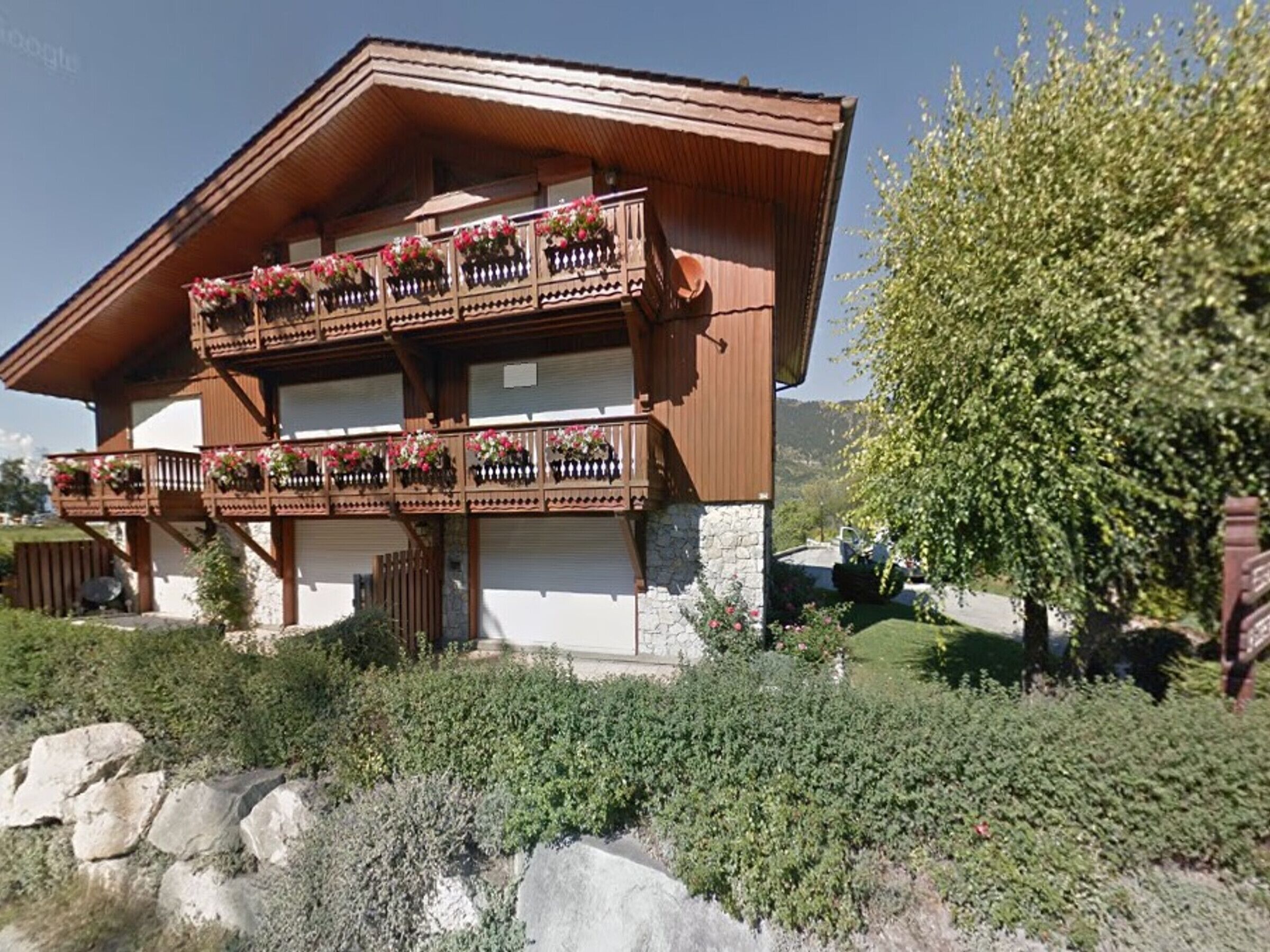 A two-story chalet with wooden balconies adorned with flower boxes, set in a mountainous area with lush greenery.