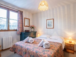 A bedroom with a bed, bedside tables, a dresser, a window with curtains, a radiator, and a hanging light fixture.