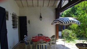 Eine Terrasse im Freien mit Steinboden, einem gedeckten Esstisch und einem gestreiften Sonnenschirm, der Schatten spendet.