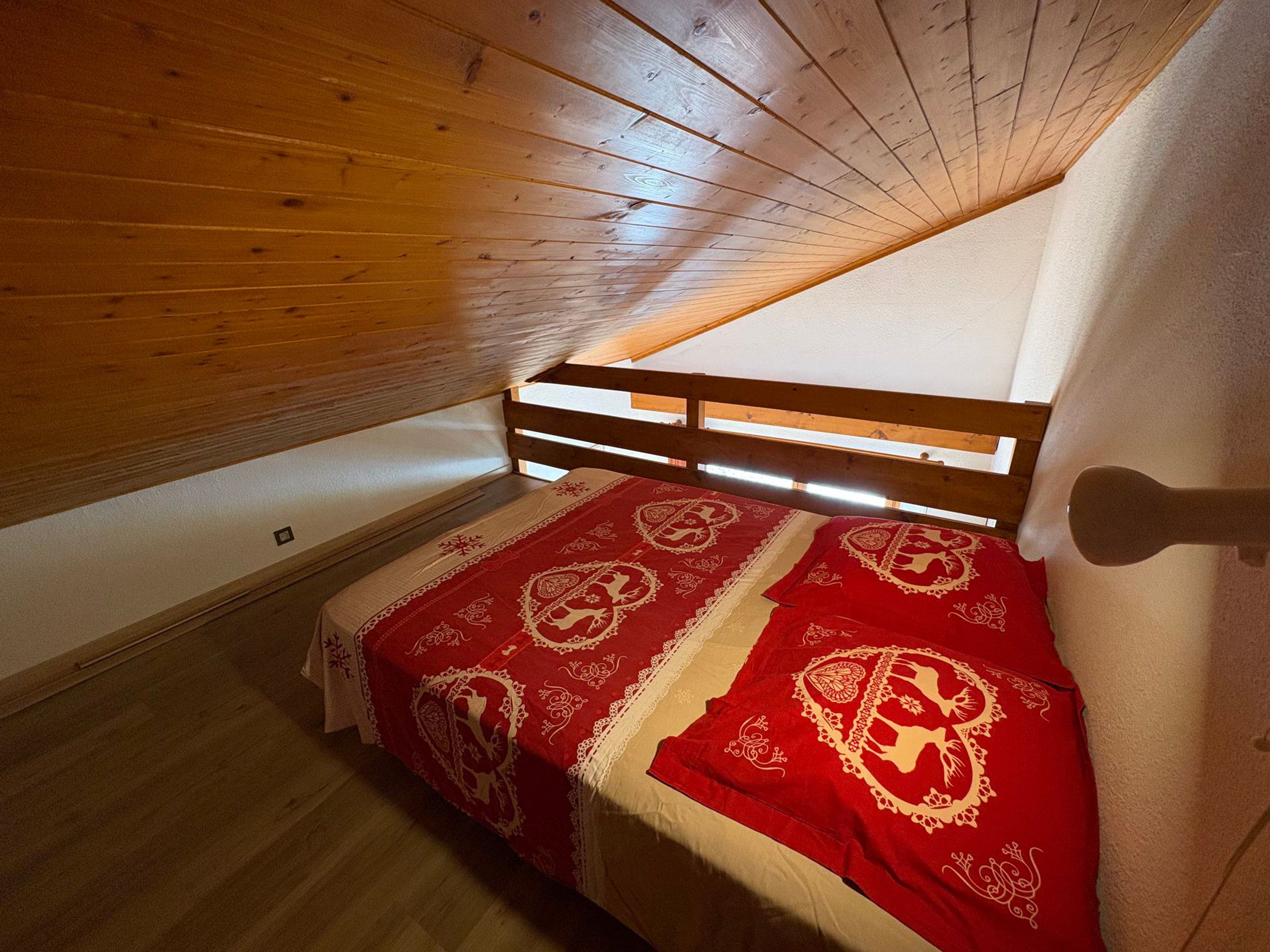 A small, cozy attic bedroom with a sloped ceiling, wooden beams, and a single bed with a red and white patterned bedspread.