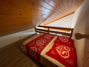 A small, cozy attic bedroom with a sloped ceiling, wooden beams, and a single bed with a red and white patterned bedspread.