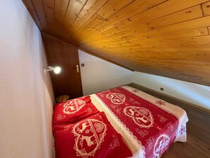 A small, cozy room with a sloped ceiling, a single bed with a red and white patterned bedspread, and a wooden door.