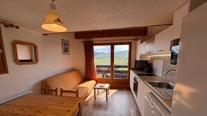 Un espacio habitable compacto con una mesa de comedor de madera, un sofá, una pequeña cocina con un fregadero y cocina, y un ventanal con vistas a un paisaje verde.