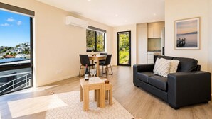 A modern living room with a black sofa, a dining table with chairs, and a large window offering a view of the sea. - View Studio | Derwent River Views | Sandy Bay (Sandy Bay)