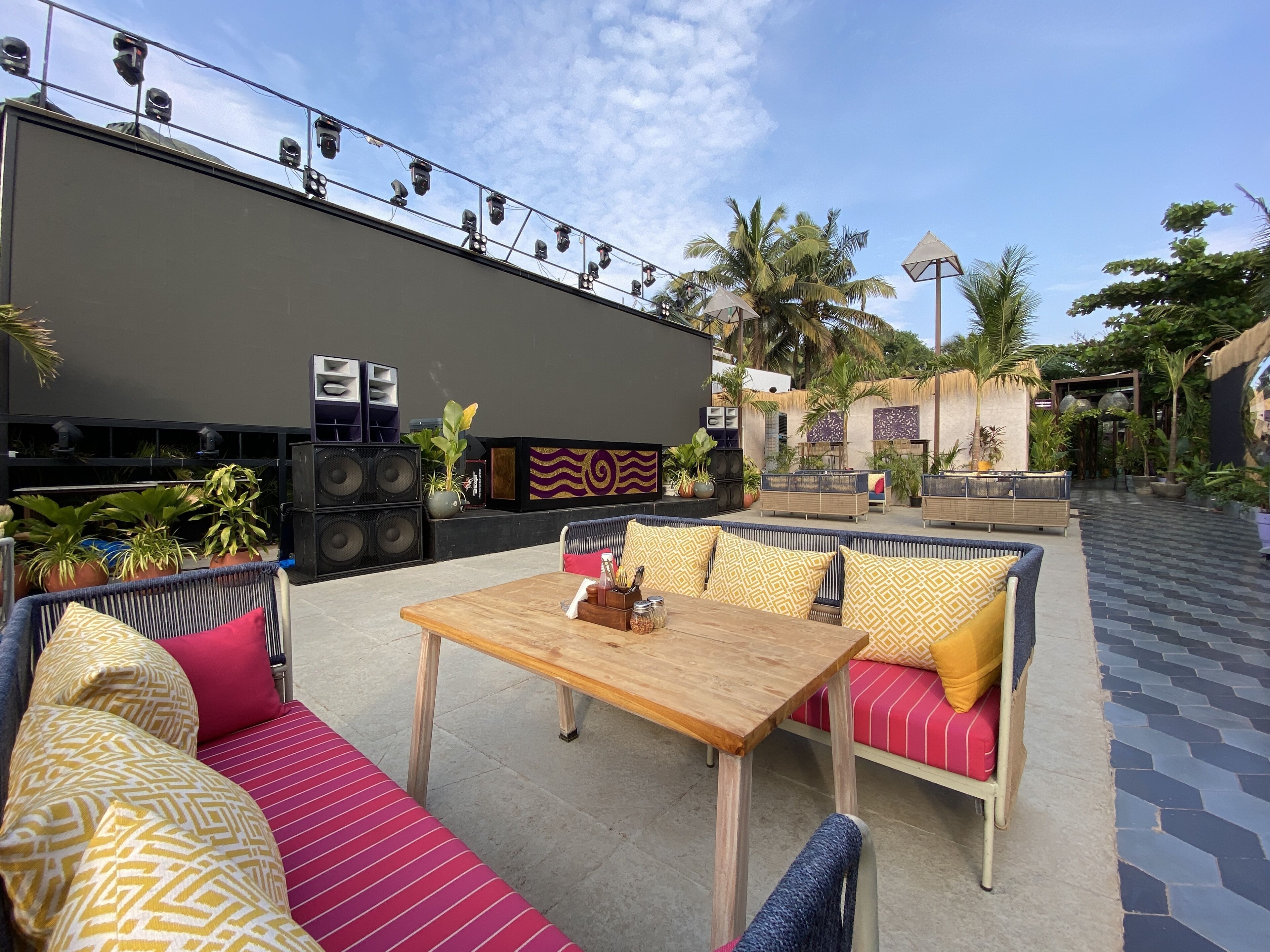 An outdoor seating area with a wooden table, cushioned sofas, and a stage with speakers.