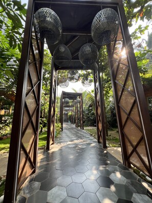 Um caminho com ladrilhos hexagonais serpenteia por uma estrutura com paredes de tela metálica e luminárias esféricas penduradas.