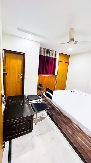 A compact room with a bed, a desk, two chairs, a wardrobe, and a ceiling fan. - Roomshala 138 Hotel Tourist palace (New Delhi)