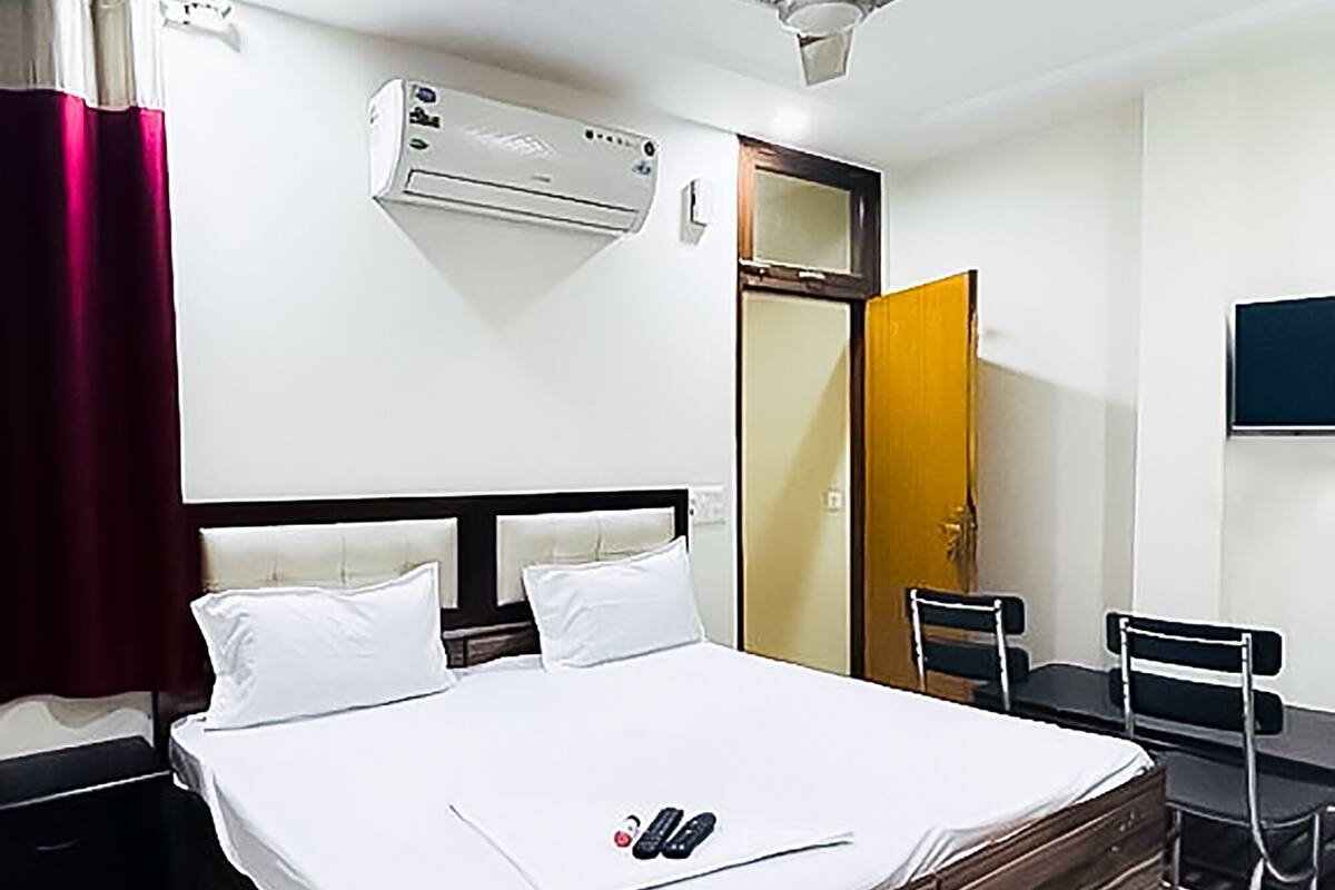 A hotel room with a bed, a desk, a chair, an air conditioning unit, a ceiling fan, and a mirror.