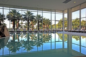 A large indoor swimming pool with clear reflections of palm trees, surrounded by lounge chairs and large windows.